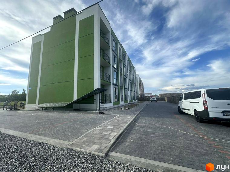 Зовнішній вигляд будинку ЖК Brickwood 2 з зеленими та білими фасадами, балконами та прибудинковою територією з бруківки та паркуванням.