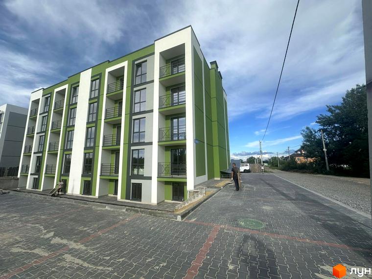 Зовнішній вигляд новобудови ЖК Brickwood 2 з зеленими та білими фасадами, балконами та паркомісцями перед будинком.