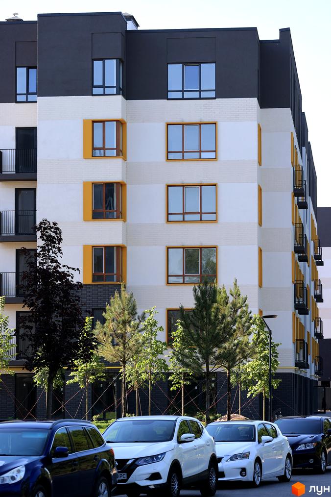 Фасад житлового комплексу Молодість з жовтими вікнами та балконами, перед ним припарковані автомобілі та зелені дерева.