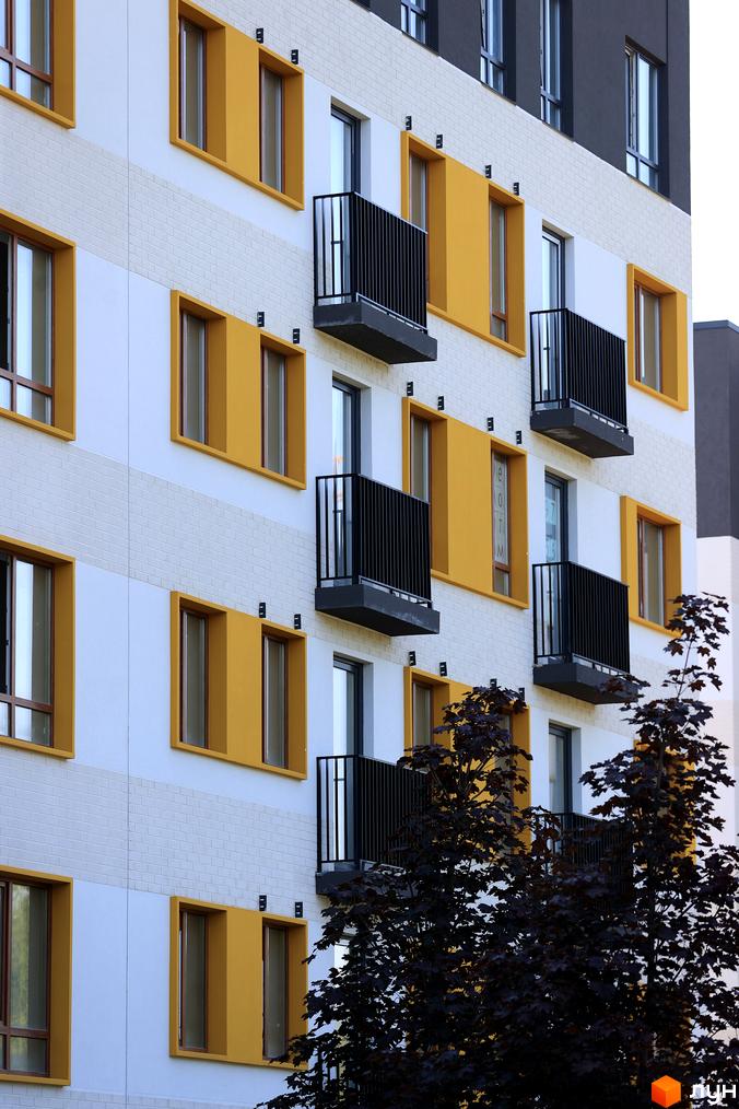 Фасад житлового комплексу Молодість з жовтими елементами та балконами. На передньому плані видно темно-зелене листя дерев.