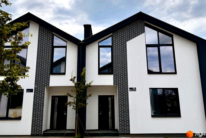 Таунхауси Idilika Home з білою штукатуркою та чорною цеглою, великими вікнами та чорними дверима. Навколо зелені дерева та хмарне небо.