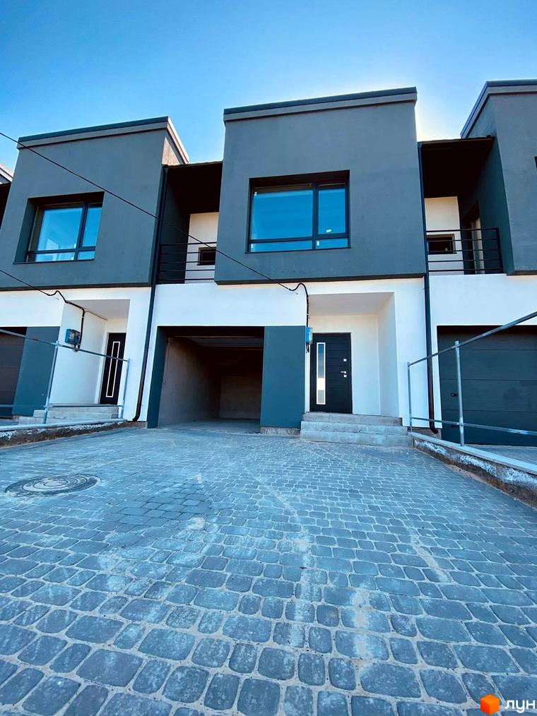 Зовнішній вигляд таунхаусів Molodizhna Town з сірими фасадами, великими вікнами та бруківкою перед будинками. Сучасна житлова забудова з гаражними воротами та входами в будинки.