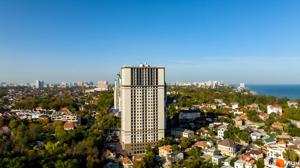 Житловий комплекс Посейдон на фоні міста та моря. Висотна будівля з великою кількістю вікон, розташована серед зелених насаджень.