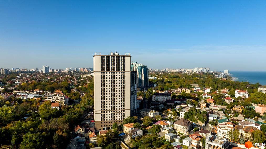 Висотна будівля ЖК Посейдон на тлі міста та моря. Архітектура будівлі з великою кількістю вікон, навколо зелені насадження та малоповерхова забудова.