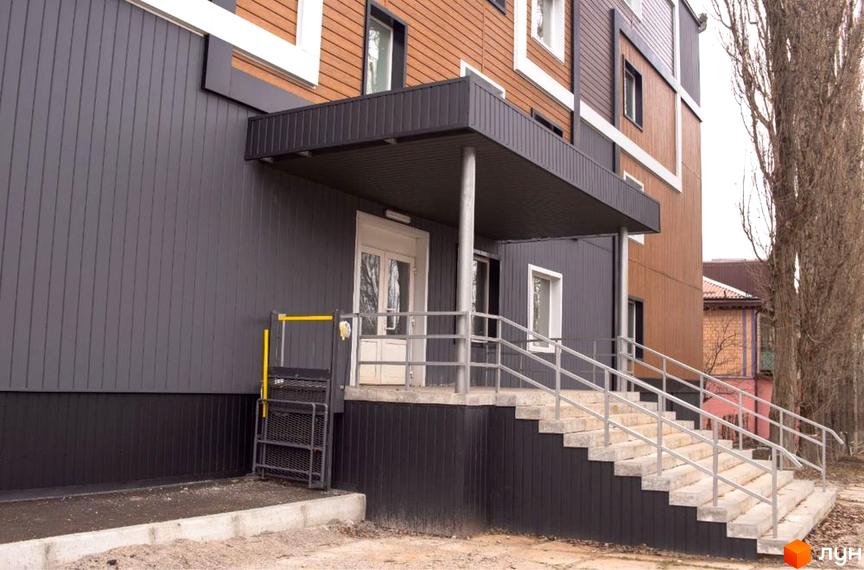 Зовнішній вигляд будинку ЖК Героїв Маріуполя з сірим фасадом, коричневими елементами та сходами з поручнями. Прибудинкова територія з під'їздом та ліфтом для людей з інвалідністю.