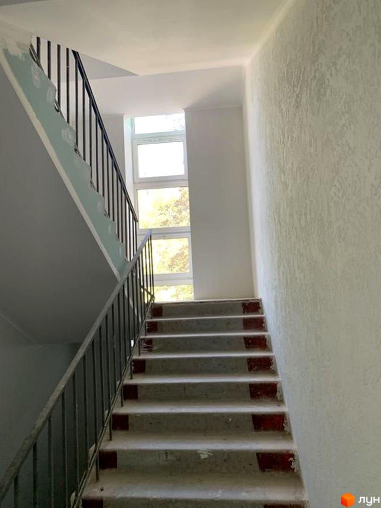 Сходи в новобудові ЖК Героїв Маріуполя. Білі стіни та сходи з металевими поручнями ведуть до вікон з видом на дерева.