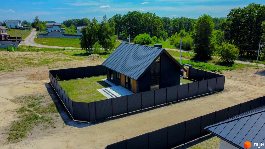 Котеджне містечко SMADE Park: чорний двоповерховий будинок з темним дахом та великими вікнами, огороджений темним парканом. Навколо будинку зелена трава та піщана дорога.