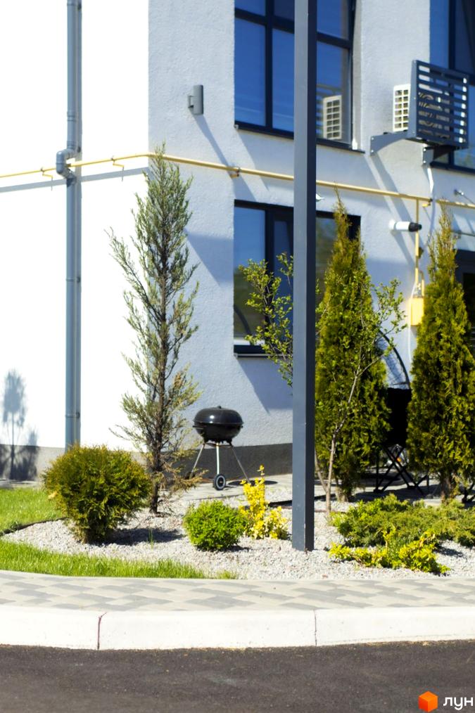 Зовнішній вигляд білої новобудови з темними вікнами та прибудинковою територією з деревами та барбекю. Таунхауси DREAM HOUSE.