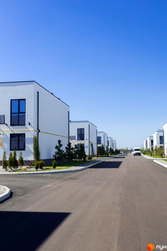 Білі таунхауси DREAM HOUSE з темними вікнами розташовані вздовж асфальтованої дороги. Прибудинкова територія озеленена газонами та деревами.