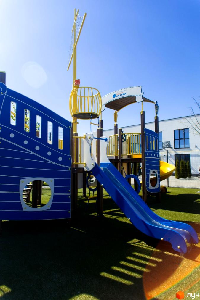 Дитячий майданчик у вигляді корабля з гіркою та елементами для лазіння. Територія комплексу DREAM HOUSE з озелененням та сучасними будівлями.