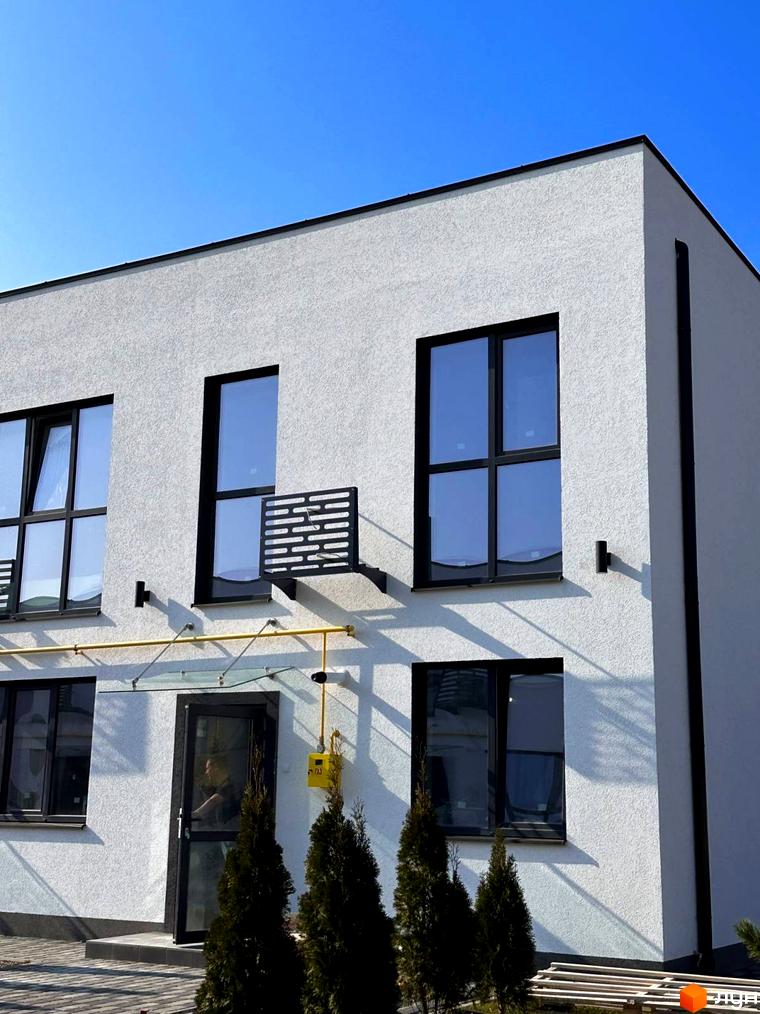 Таунхауси DREAM HOUSE: білий фасад будинку з чорними вікнами та скляним навісом над входом. Перед будинком ростуть хвойні дерева.