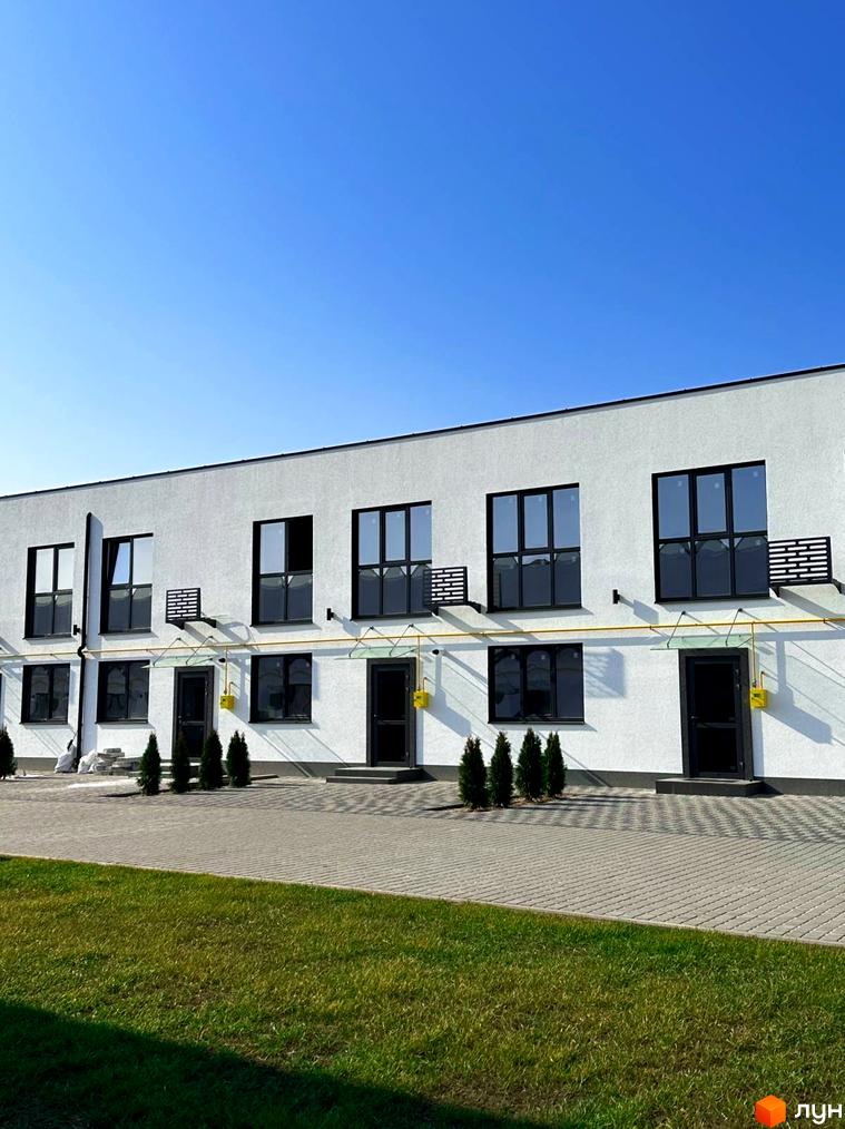 Ряд білих таунхаусів DREAM HOUSE з чорними вікнами та дверима, жовтими газовими трубами. Перед будинками бруківка та зелений газон під чистим небом.