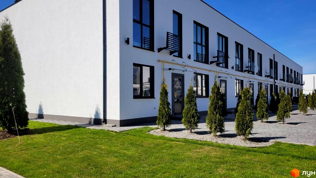 Зовнішній вигляд білих таунхаусів DREAM HOUSE з чорними вікнами та дверима, зелений газон та доглянуті туї перед будинками.