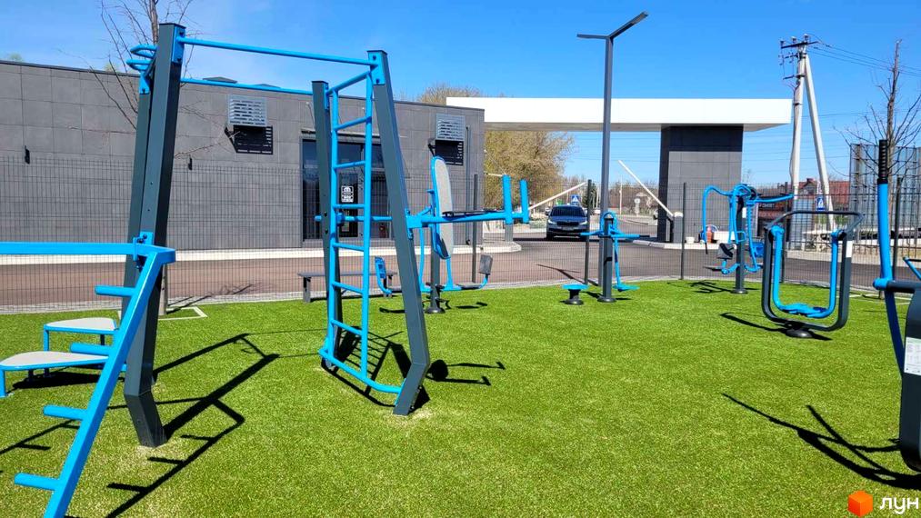 Спортивний майданчик з тренажерами біля будівель житлового комплексу DREAM HOUSE. Зелена трава та сині тренажери створюють привабливий простір для відпочинку.