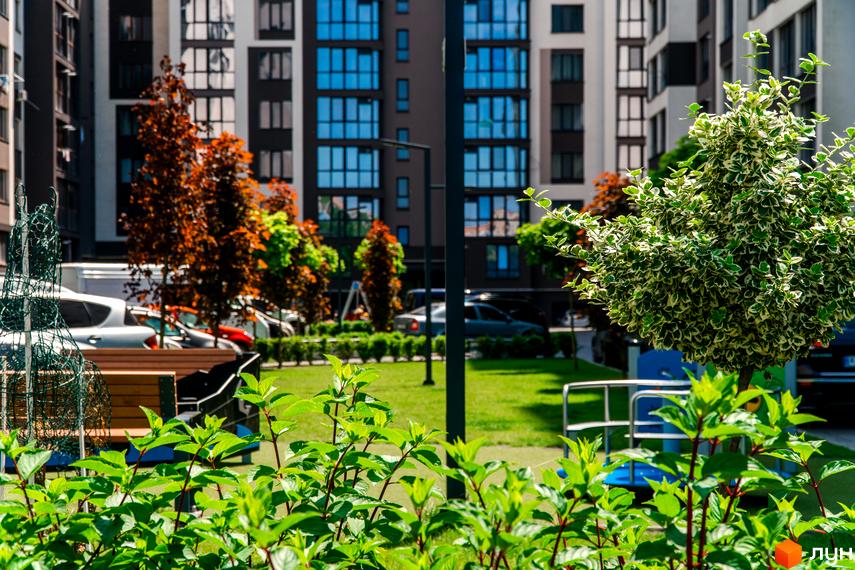 Зображення двору Житлового комплексу Паркова Алея. На фото будівля та зелена прибудинкова територія з деревами, кущами та лавками.