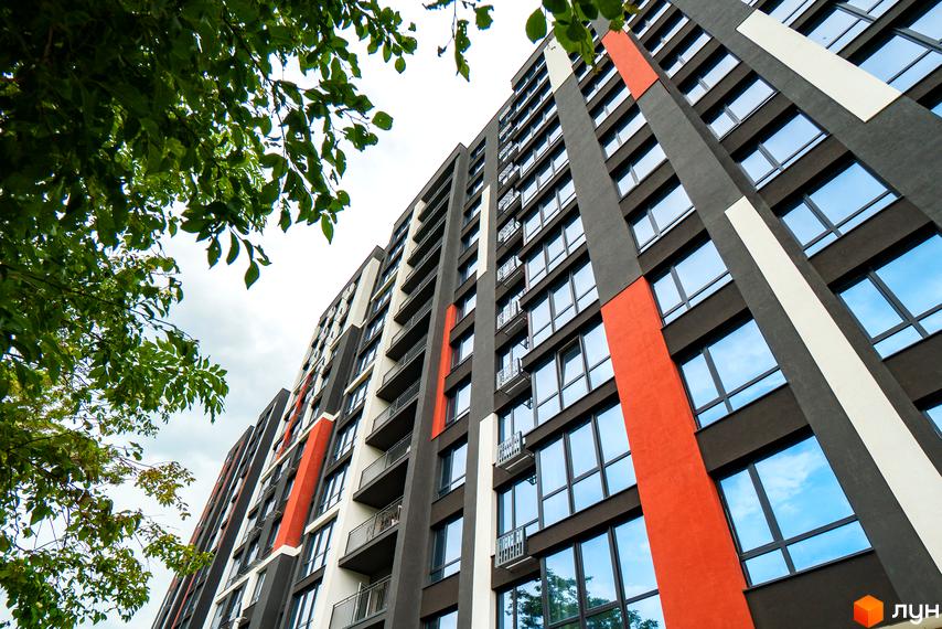 Висотна будівля ЖК U One з великими вікнами та балконами, фасад пофарбований у сірий, білий та помаранчевий кольори. Зелені гілки дерев обрамляють кут будинку.