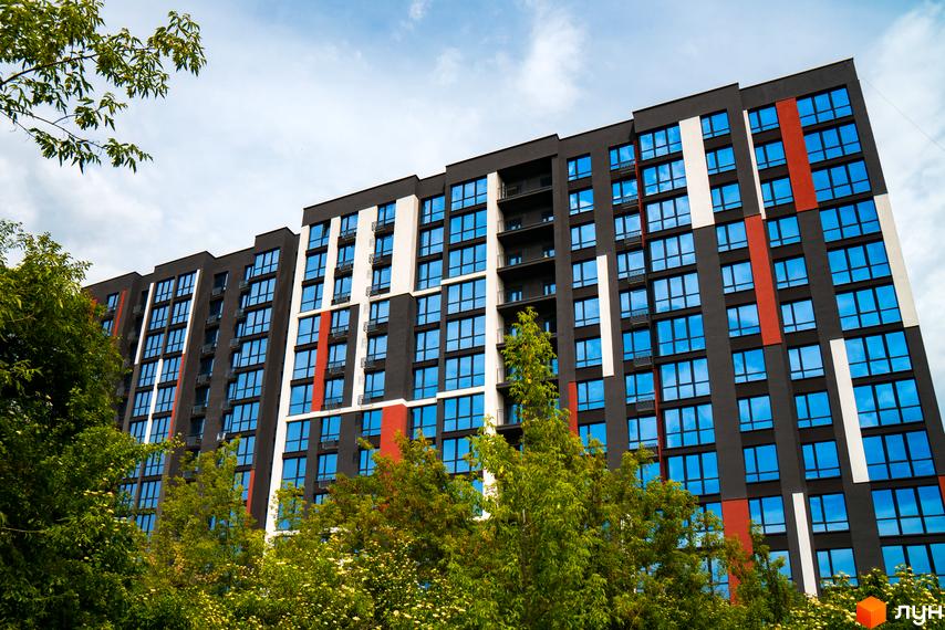 Висотна будівля ЖК U One з великою кількістю вікон та балконів, фасади пофарбовані у різні кольори. Перед будинком зелені дерева.