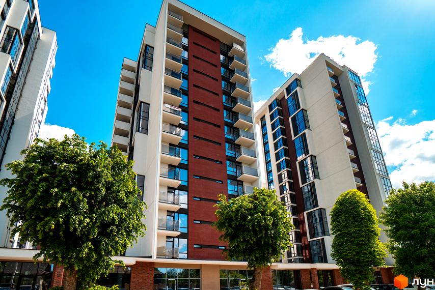 Житловий район Manhattan — високі будинки біло-червоного кольору з численними балконами та вікнами. Перед будівлями ростуть зелені дерева під яскравим небом.