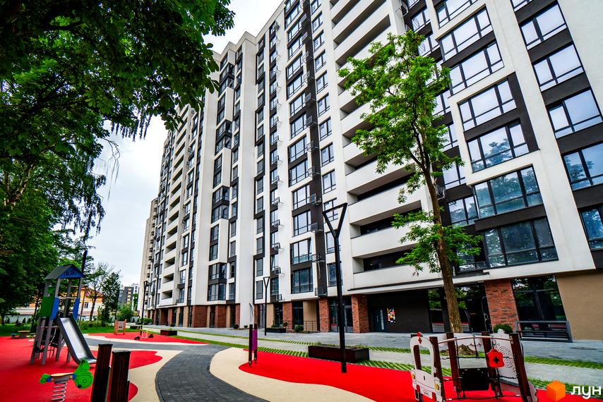 Житловий район Manhattan: висотна біла новобудова з великими темними вікнами та балконами. Поруч дитячий майданчик з червоним покриттям та зелені дерева.