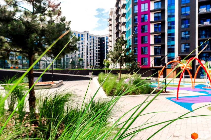 Вигляд на будівлю Житлового комплексу Family Plaza та дитячий майданчик. На передньому плані зелені насадження.