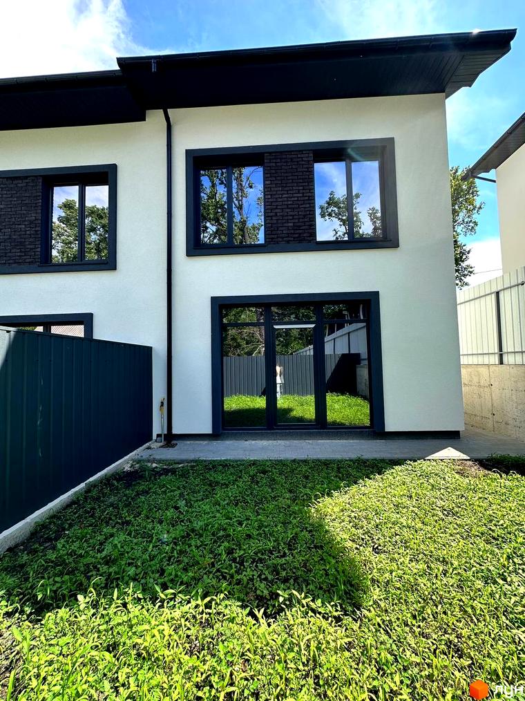 Білий двоповерховий будинок з темними вікнами та дверима. Перед будинком газон з зеленою травою, а також зелений паркан.