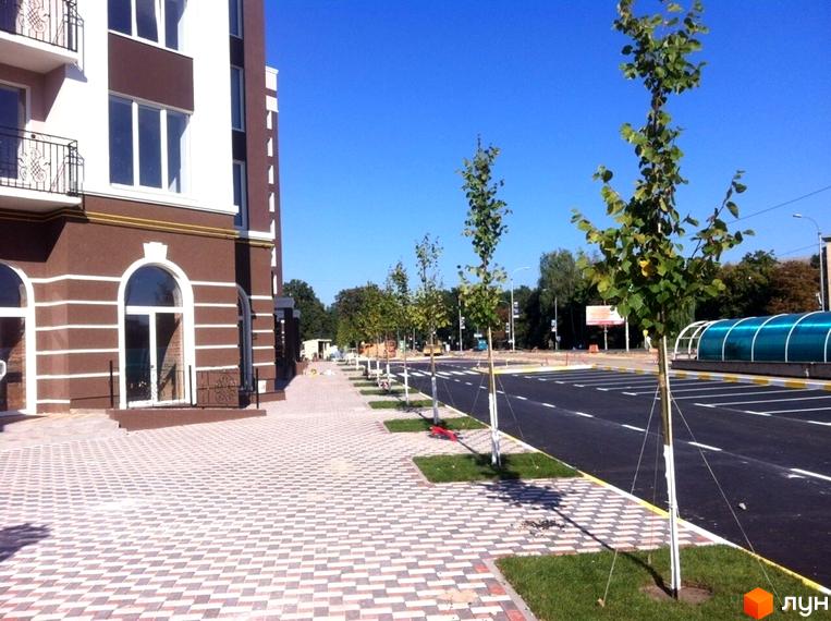 Новобудова ЖК Центральний з коричнево-білим фасадом та арковими вікнами. Прибудинкова територія з бруківкою, зеленими насадженнями та дорогою.