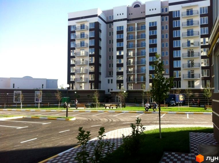 Зовнішній вигляд багатоповерхового будинку ЖК Центральний з балконами та вікнами. Прибудинкова територія з парковкою, лавками та деревами.