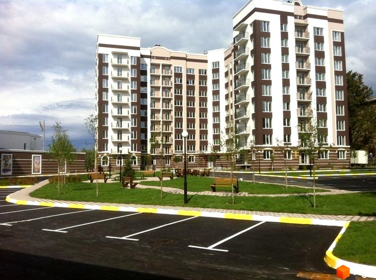 Житловий комплекс Центральний з коричнево-білим фасадом та балконами. Біля будинку розташований паркінг, зелена зона з лавками та деревами.