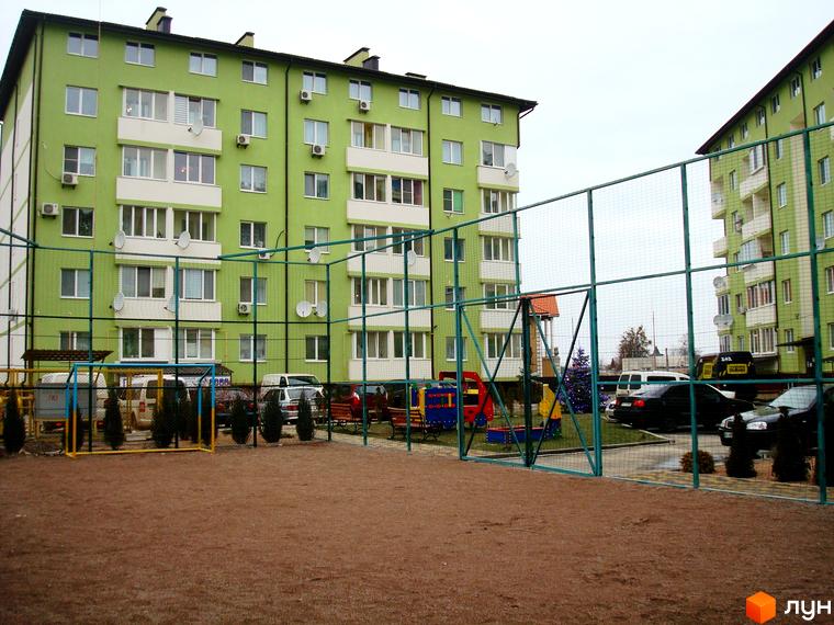 Зовнішній вигляд зелених багатоповерхових будинків ЖК Смарагдове місто. На прибудинковій території розташований дитячий майданчик та спортивний майданчик, огороджений сіткою.