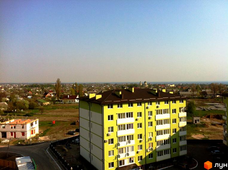 Зовнішній вигляд яскраво-жовтої новобудови ЖК Смарагдове місто з білими вікнами та балконами. Навколо зелена територія з іншими будинками та дитячим майданчиком.