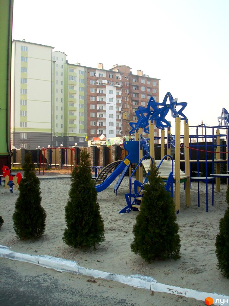 Новобудова ЖК Смарагдове місто у Нових Петрівцях. Сучасний дитячий майданчик з гірками та гойдалками на піщаному покритті поруч із зеленими туями.