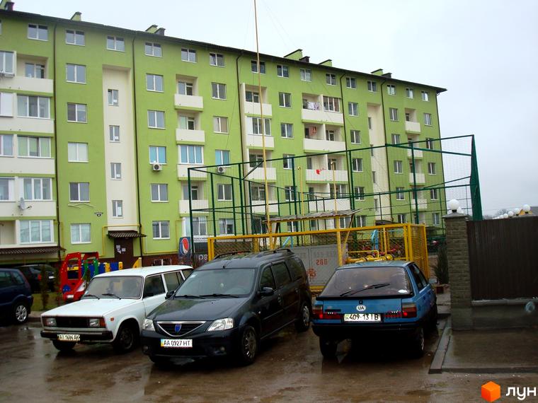 Житловий комплекс Смарагдове місто у Нових Петрівцях. Зелена фасадна частина багатоповерхового будинку з білими балконами та вікнами, прибудинкова територія з автомобілями.