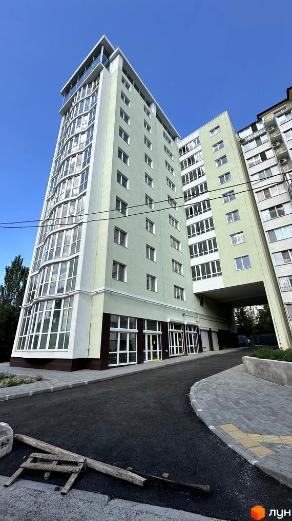 Зовнішній вигляд ЖК Потьомкінський з вигнутим фасадом та великою кількістю вікон. Прибудинкова територія з асфальтованою дорогою та бруківкою.