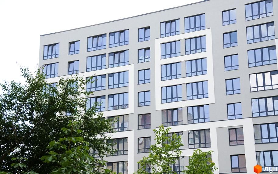 Новобудова Молодіжний з великою кількістю вікон та зеленими деревами на передньому плані. Зовнішній вигляд фасаду будинку з сірими та білими елементами.