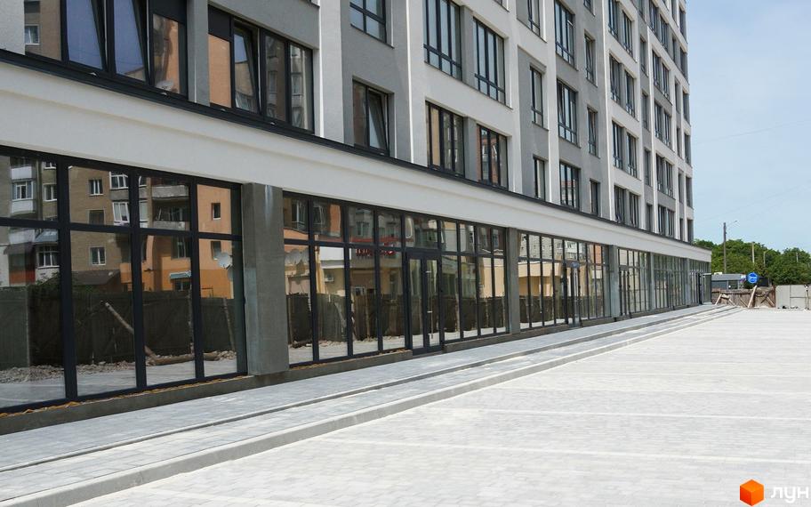 Зовнішній вигляд новобудови ЖК Молодіжний з великими вікнами та паркуванням. Будинок має сіро-білий фасад та відображає навколишні будівлі.