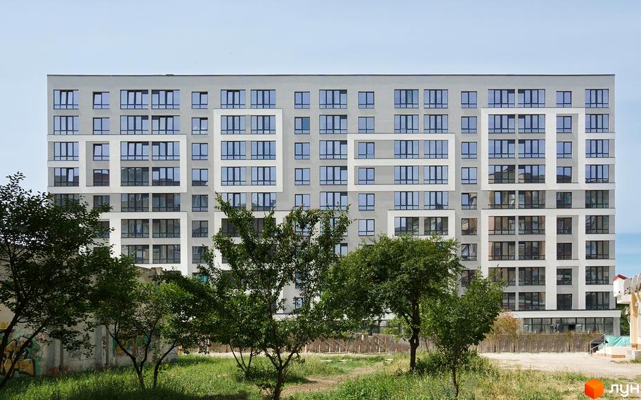Зовнішній вигляд будинку ЖК Молодіжний з численними вікнами та сірою фасадною частиною. На передньому плані зелені дерева та невисока огорожа.
