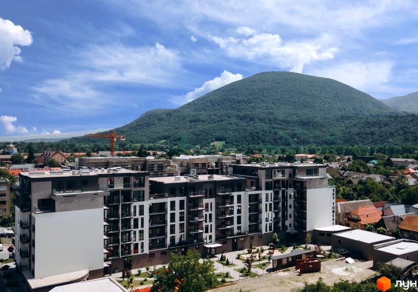 Висотна будівля ЖК Sky Towers з білими та сірими фасадами на фоні гірського пейзажу. На території комплексу є зелені насадження та парковка.