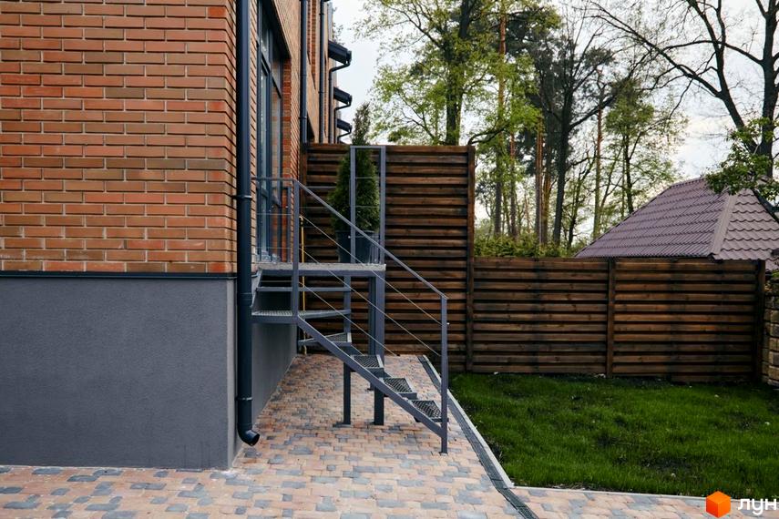 Вигляд фасаду будинку з червоної цегли та металеві сходи в Котеджному містечку Positano Park. Подвір'я з газоном та дерев'яний паркан.