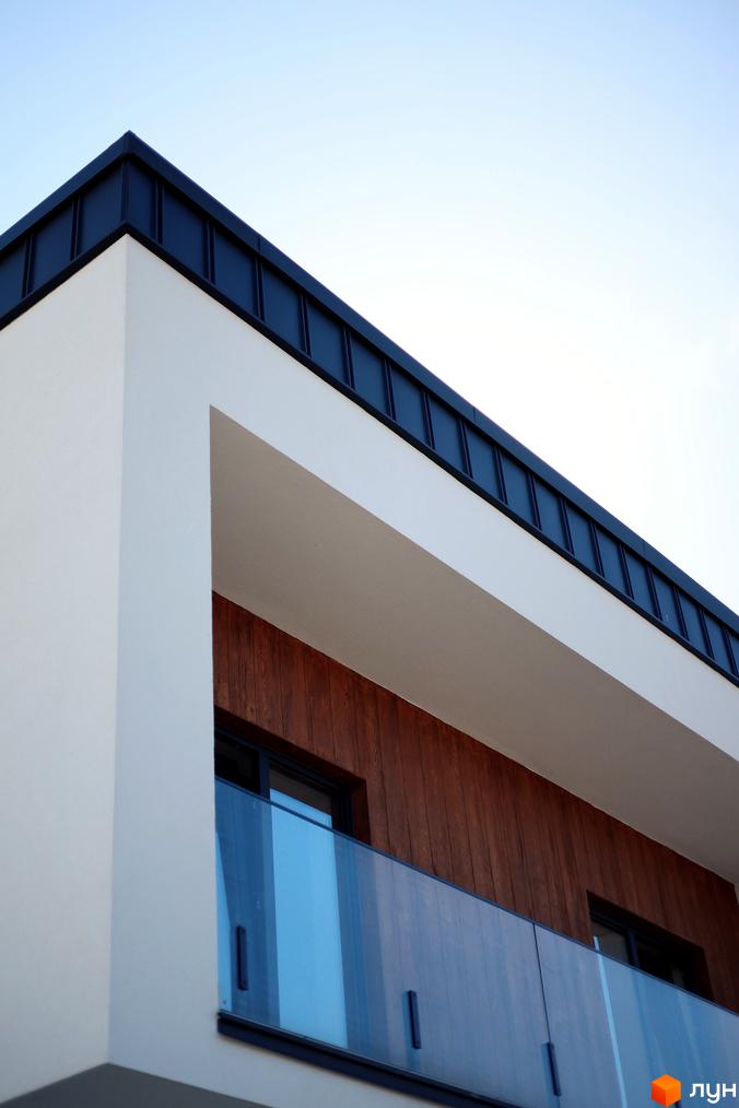 Кутовий фасад будинку з білими стінами, дерев'яною обшивкою та скляним балконом. Чорний дах з чіткими лініями на тлі блакитного неба.