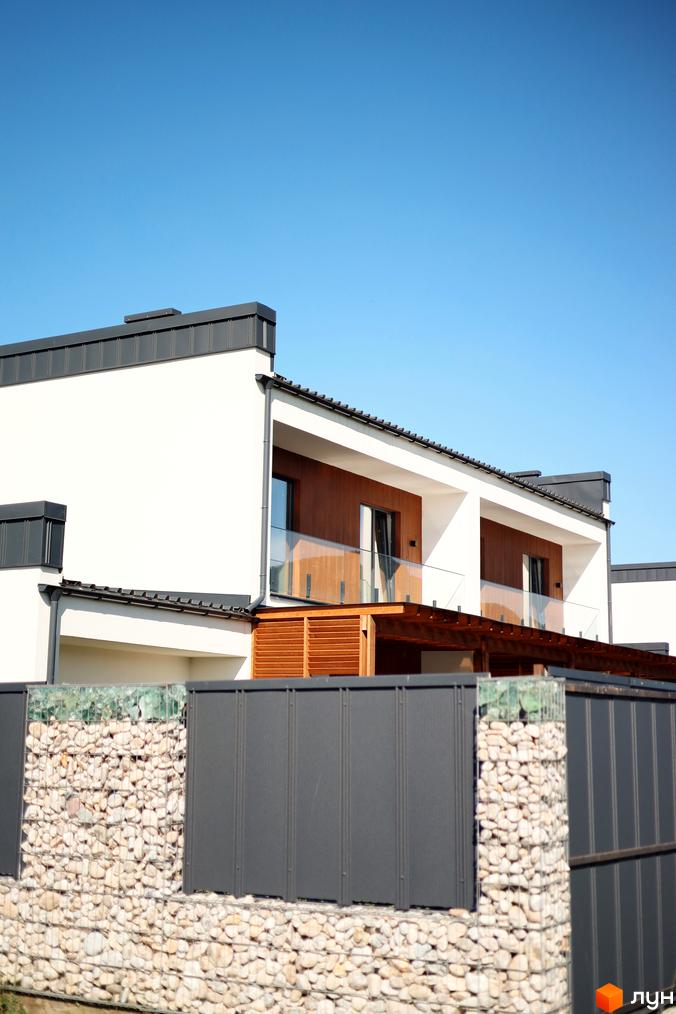 Фасад двоповерхового будинку з білими стінами та темною покрівлею. Перед будинком кам'яний паркан та зелені насадження.