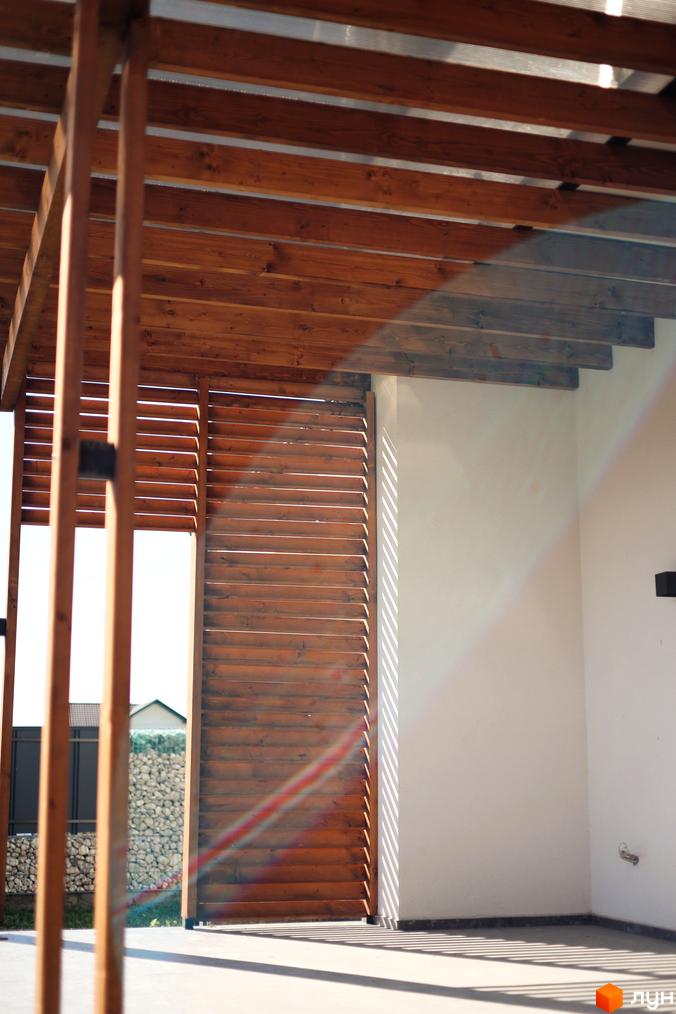 Дерев'яний навіс над терасою з ламелями та білою стіною. На задньому плані видно частину огорожі та будинки.