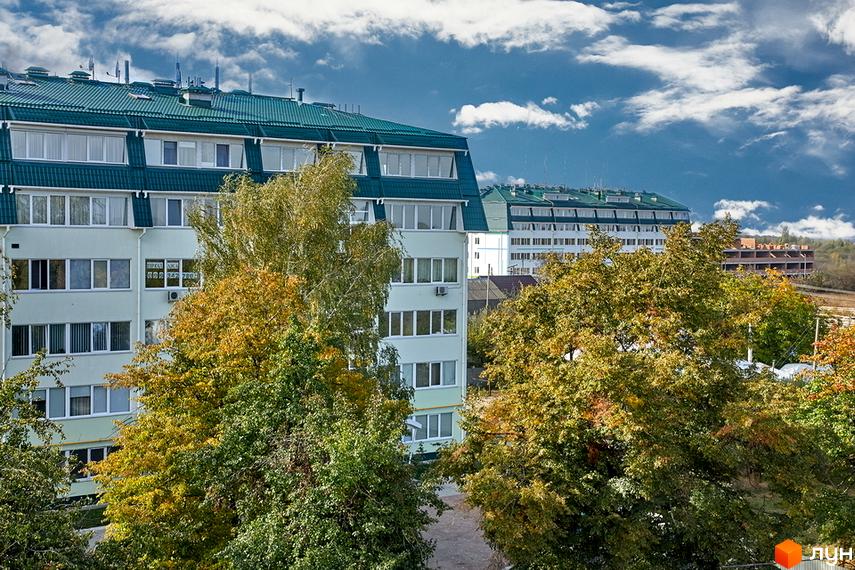 Зовнішній вигляд багатоповерхового будинку з зеленим дахом та вікнами. Навколо ростуть дерева з жовтим та зеленим листям. Комплекс