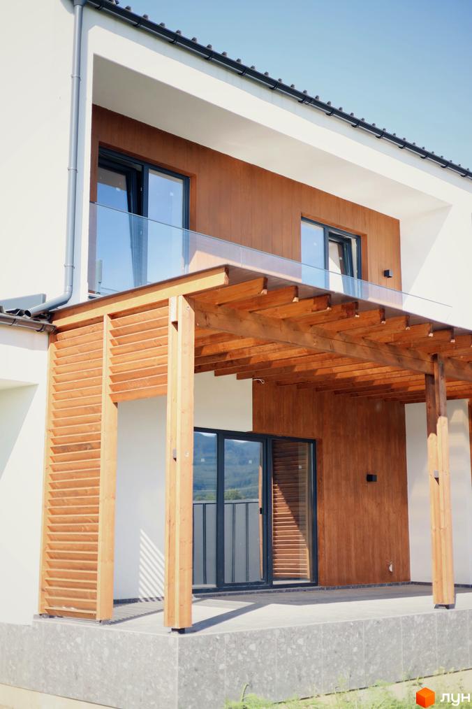 Двоповерховий будинок з дерев'яним декором та терасою. Фасад будинку виконаний у світлих тонах з великими вікнами.