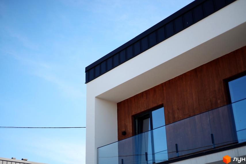 Фасад двоповерхового будинку з дерев'яним оздобленням та скляним балконом. Небо блакитне, сонячний день.