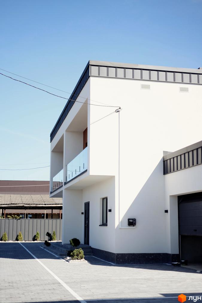 Білий двоповерховий будинок з балконами та скляними перилами. Перед будинком бруківка, газон з кущами та невеликі дерева.