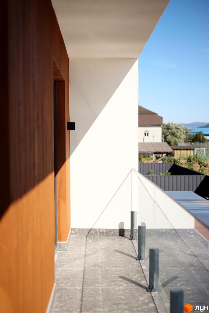 Вид на балкон житлового будинку. Скляне огородження, плитка на підлозі, білі стіни та дерев'яні панелі фасаду. На задньому плані видно інші будинки та блакитне небо.