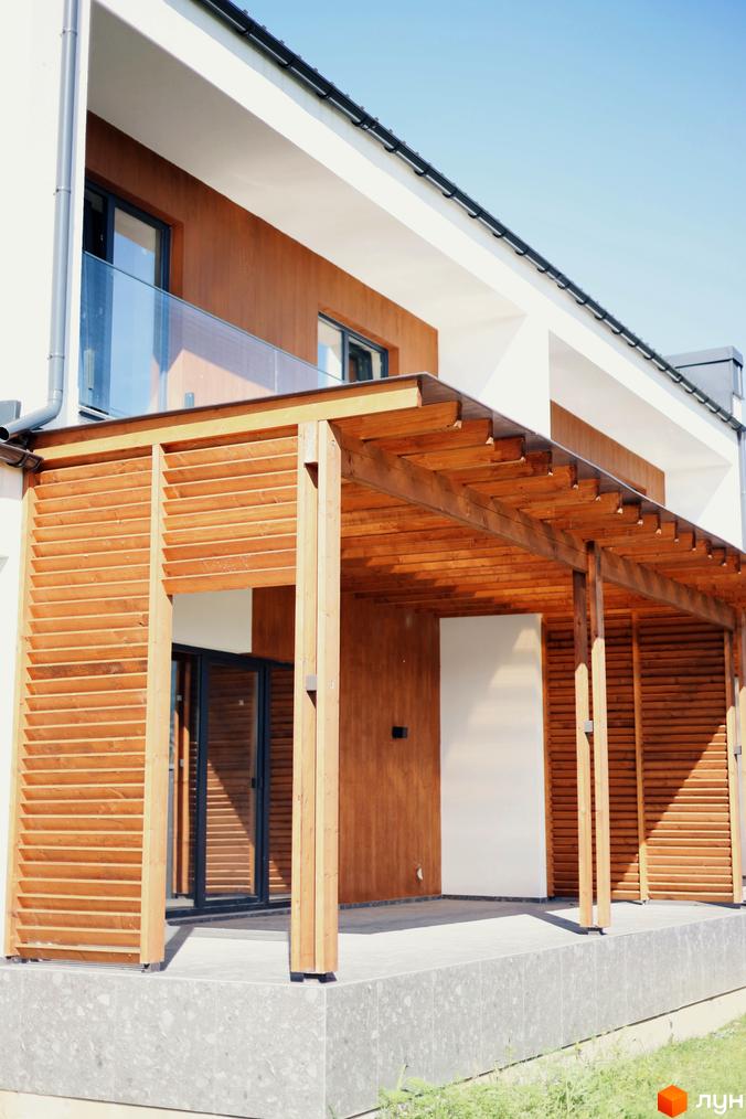 Двоповерховий будинок з дерев'яною терасою та великими вікнами. На передньому плані бетонна плита та газон.