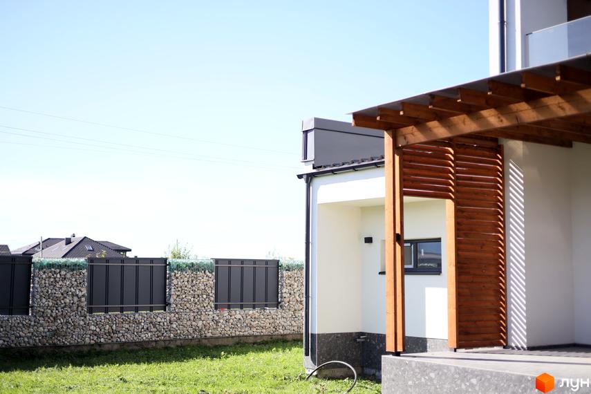 Зовнішній вигляд будинку в житловому комплексі Twins House з дерев'яною терасою та кам'яним парканом. Зелена прибудинкова територія.