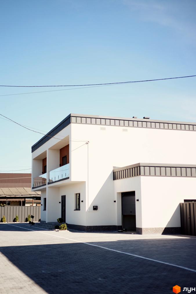 Біла будівля Twins House з балконами та гаражем, вимощена бруківкою прибудинкова територія, блакитне небо.