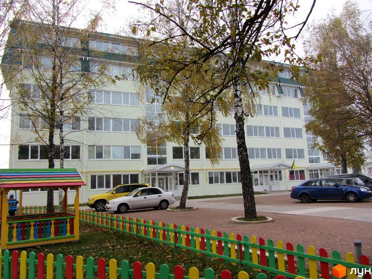 Новобудова ЖК Милі квартири з багатьма вікнами та зеленою покрівлею. Прибудинкова територія з дитячим майданчиком та автомобілями.